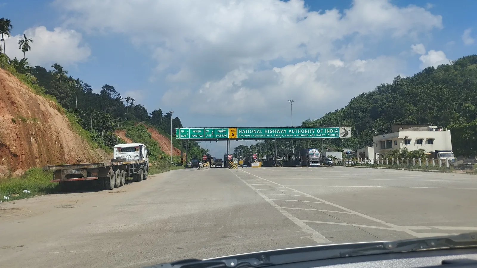 A National Highways Authority of India toll plaza seen through a car windscreen, with a green gantry sign marking FASTag lanes and trucks queuing at the booths between green hills