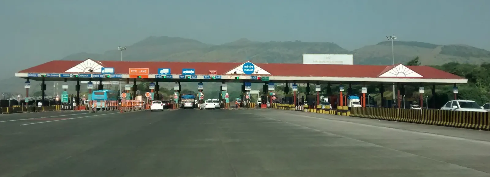 The red-roofed Talegaon toll plaza on the Mumbai-Pune Expressway with a row of booths, an orange sign marking the ETC lane on the far left and cars queuing under hazy hills