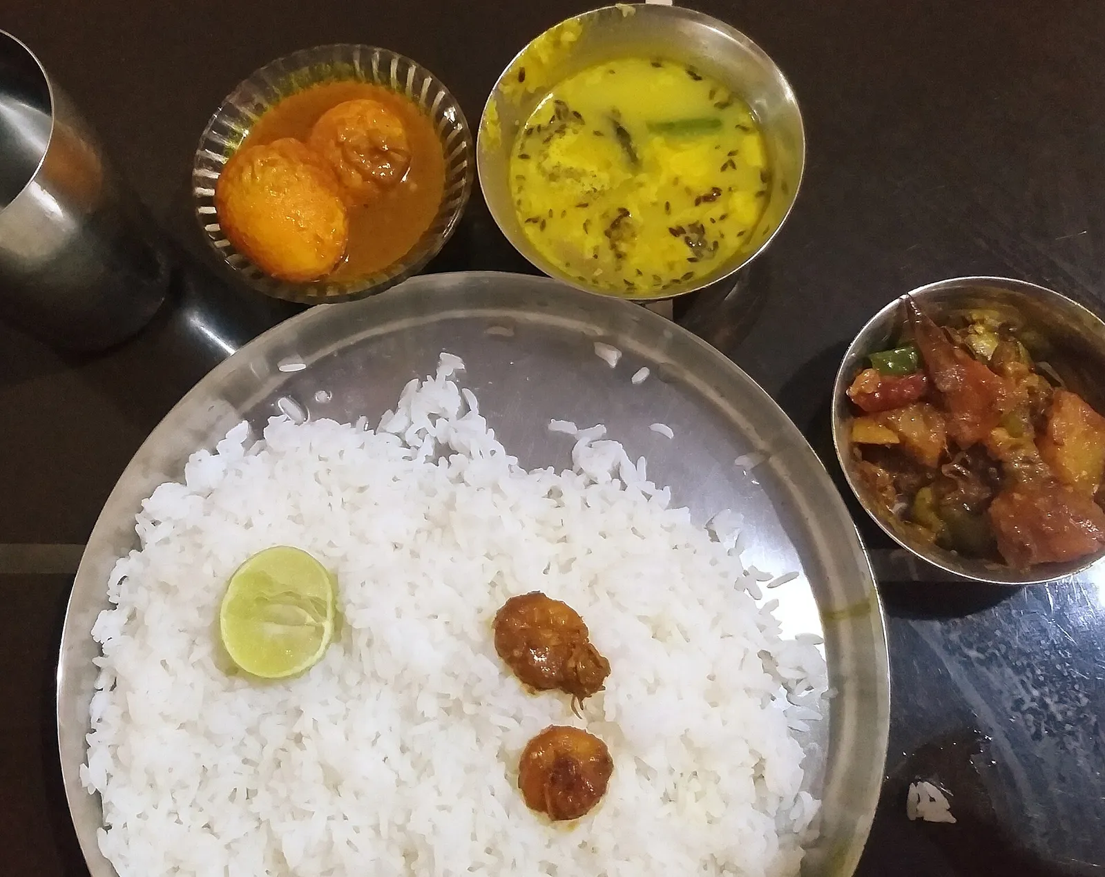 A Bengali home meal of white rice on a steel plate with bowls of yellow dal, egg curry and a mixed vegetable curry