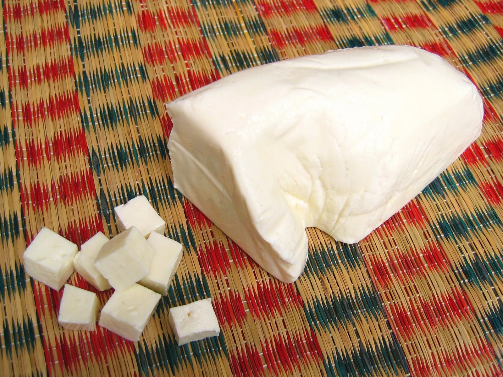 A block of fresh white paneer with several neatly cut cubes on a woven red and green mat