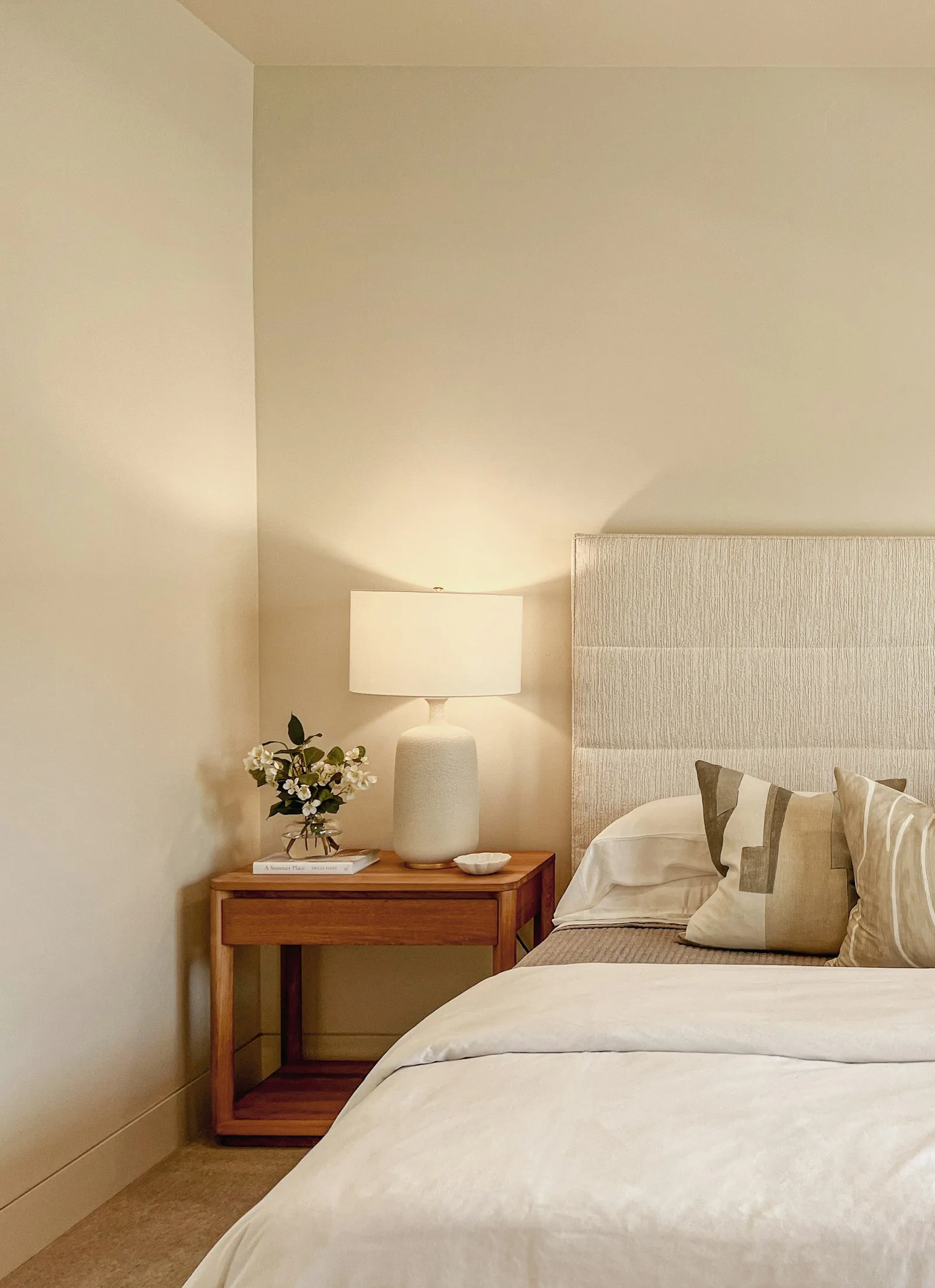 A modern bedroom with a made bed, wooden headboard, and a lit bedside lamp on a mid-century nightstand