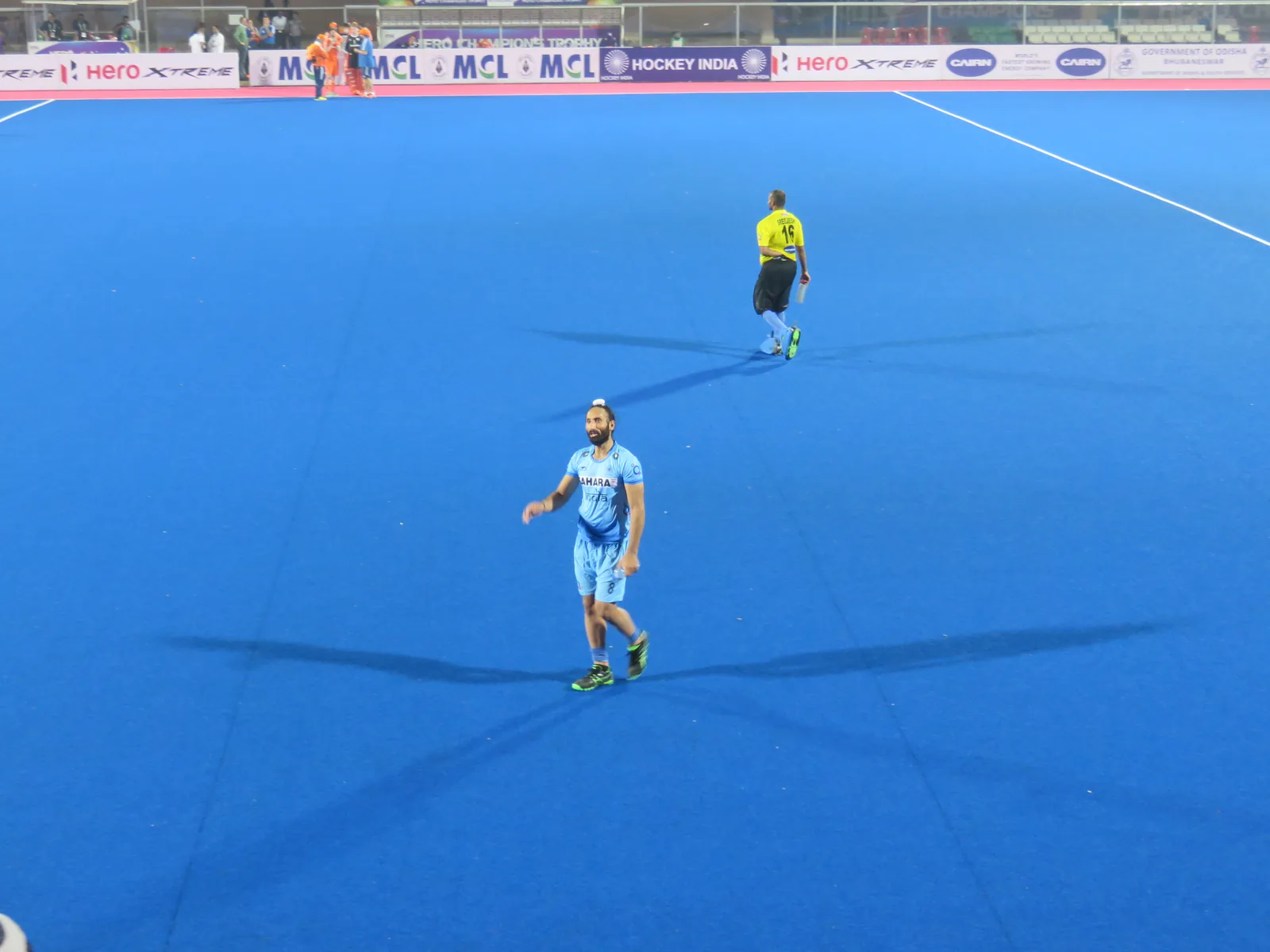 Indian hockey captain Sardar Singh in an orange jersey standing on the turf at Kalinga Stadium, holding a stick, with empty blue stands behind him