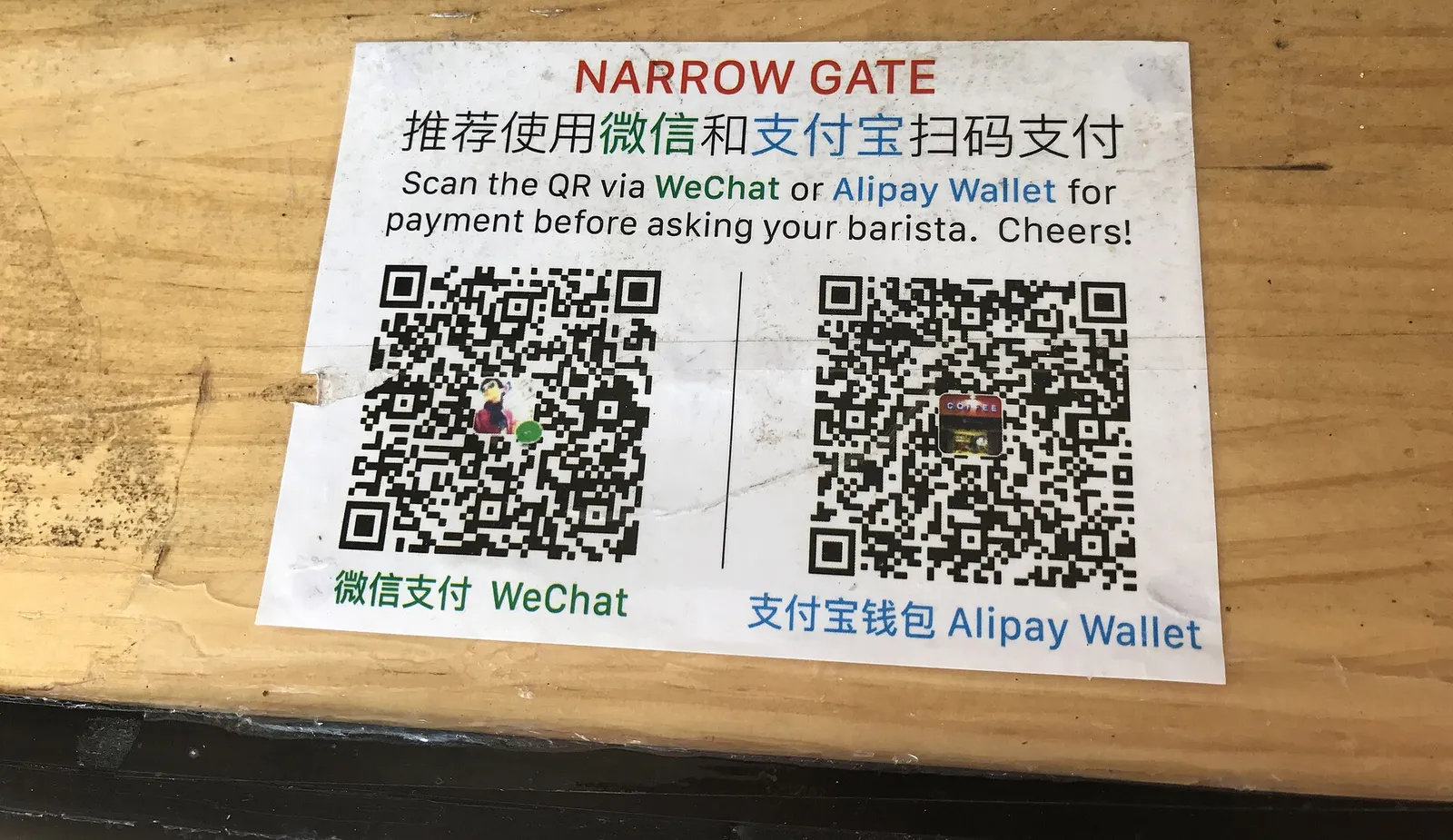 Printed sign on a wooden café counter with two QR codes, one for WeChat Pay and one for Alipay, and instructions in Chinese and English