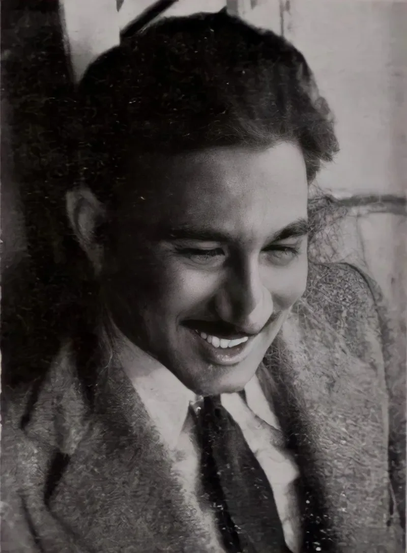 Black and white portrait of a young, smiling Khwaja Ahmad Abbas in a jacket and tie, looking down and to one side