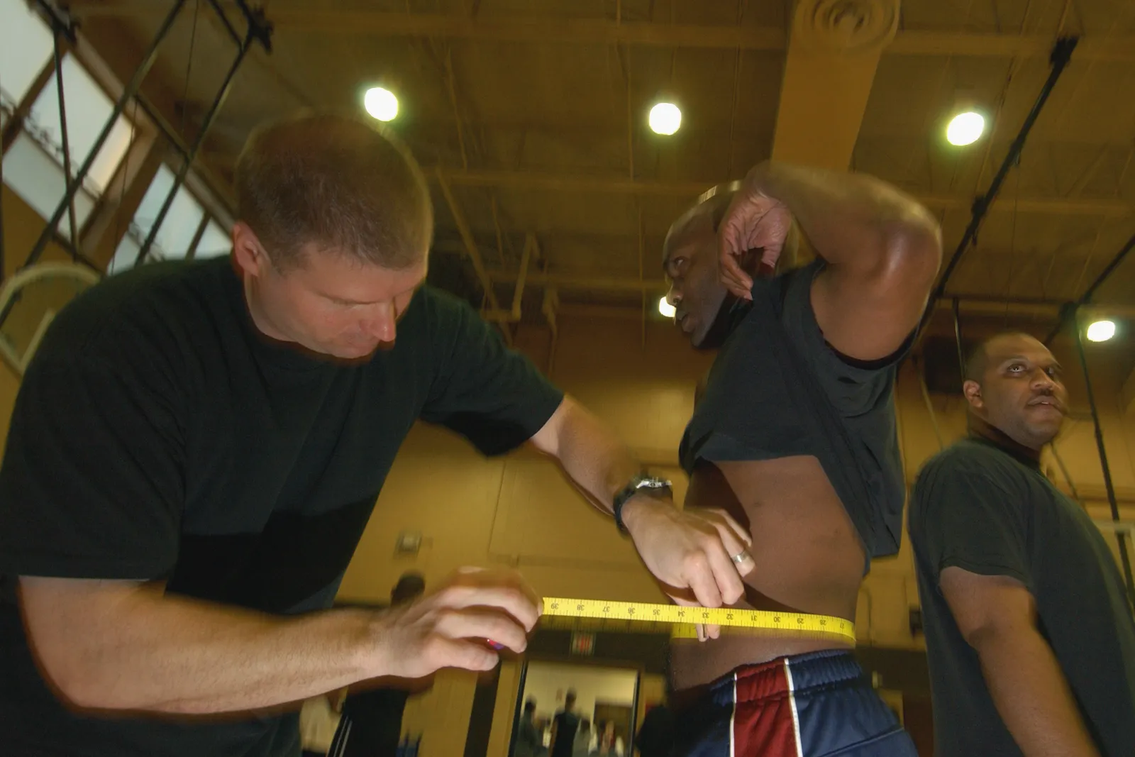 A fitness instructor wraps a yellow measuring tape around a man's bare waist in a gym