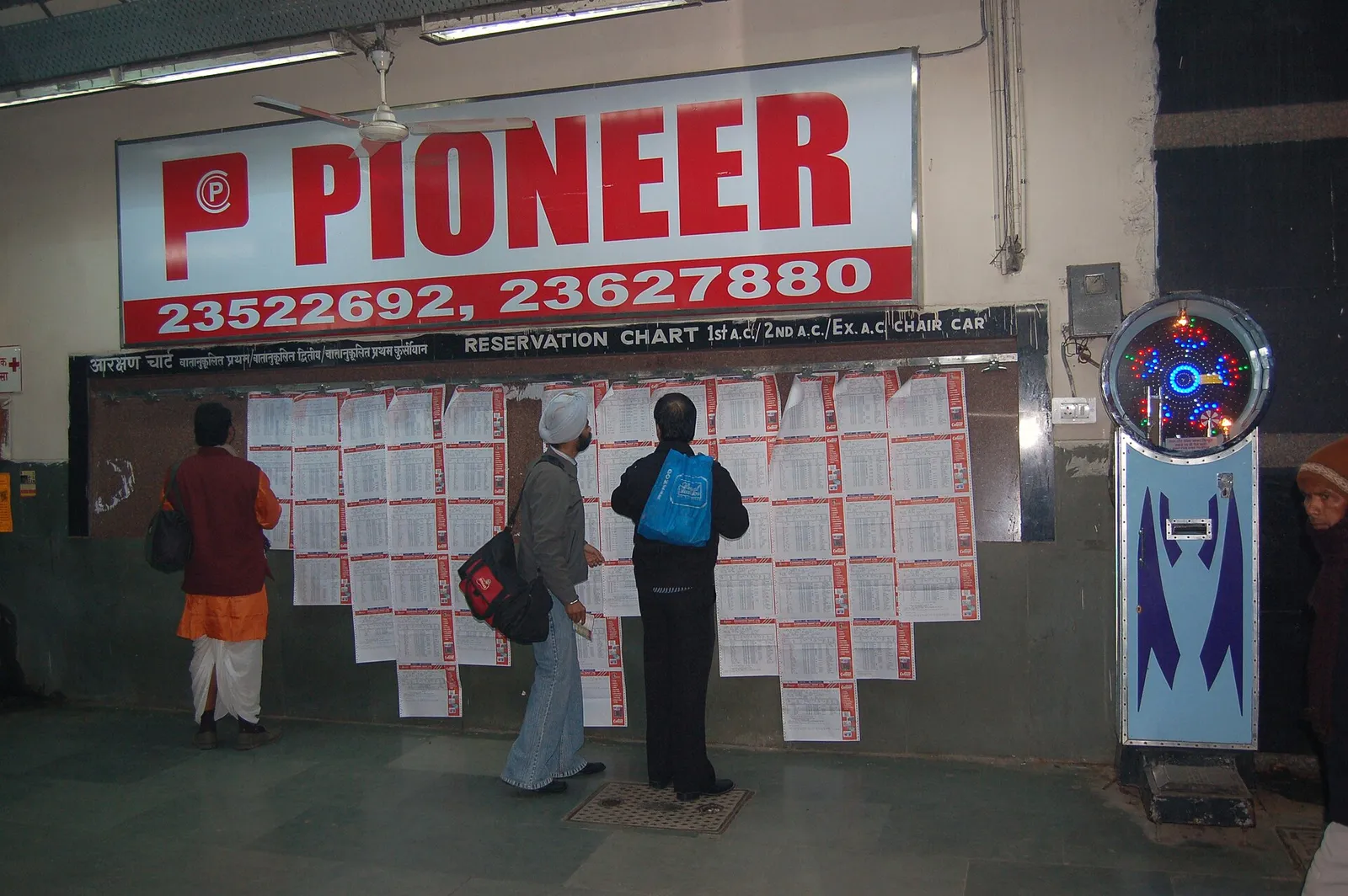 Three passengers in winter clothes reading printed reservation charts pinned in rows on a board labelled Reservation Chart 1st AC, 2nd AC, Ex AC Chair Car on a platform at New Delhi railway station