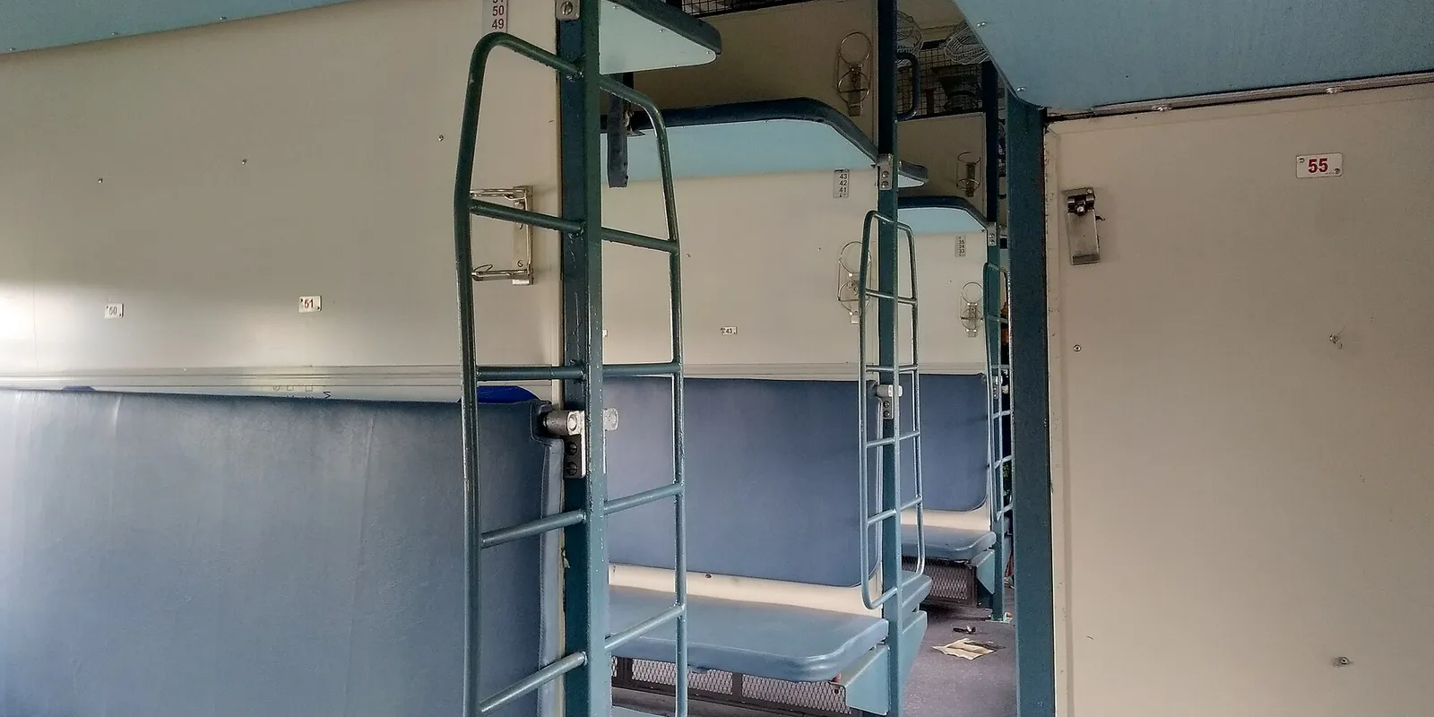 The empty interior of an Indian Railways Sleeper class coach with blue-grey berths stacked in three tiers, green metal ladders and a berth number plate reading 55