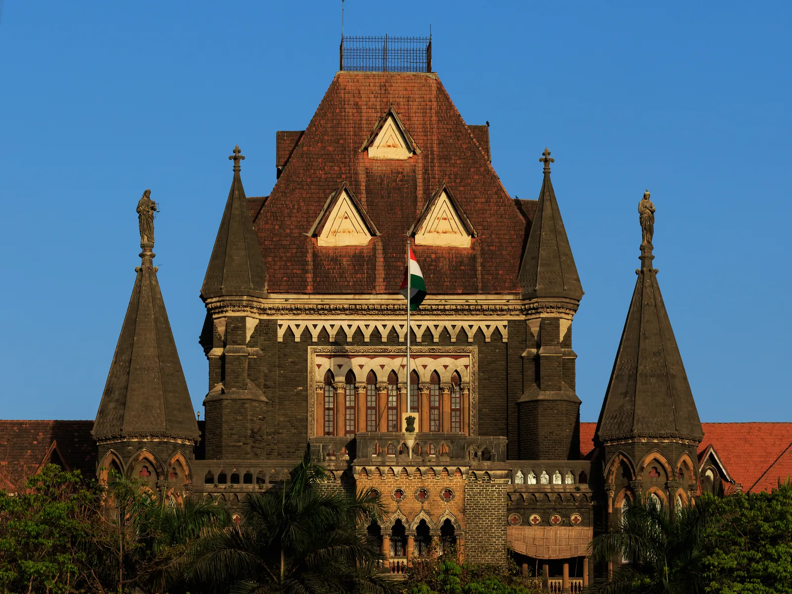 The dark stone Gothic Revival tower of the Bombay High Court, with steep red tiled gables, two pointed turrets topped by statues and an Indian flag, against a clear blue sky