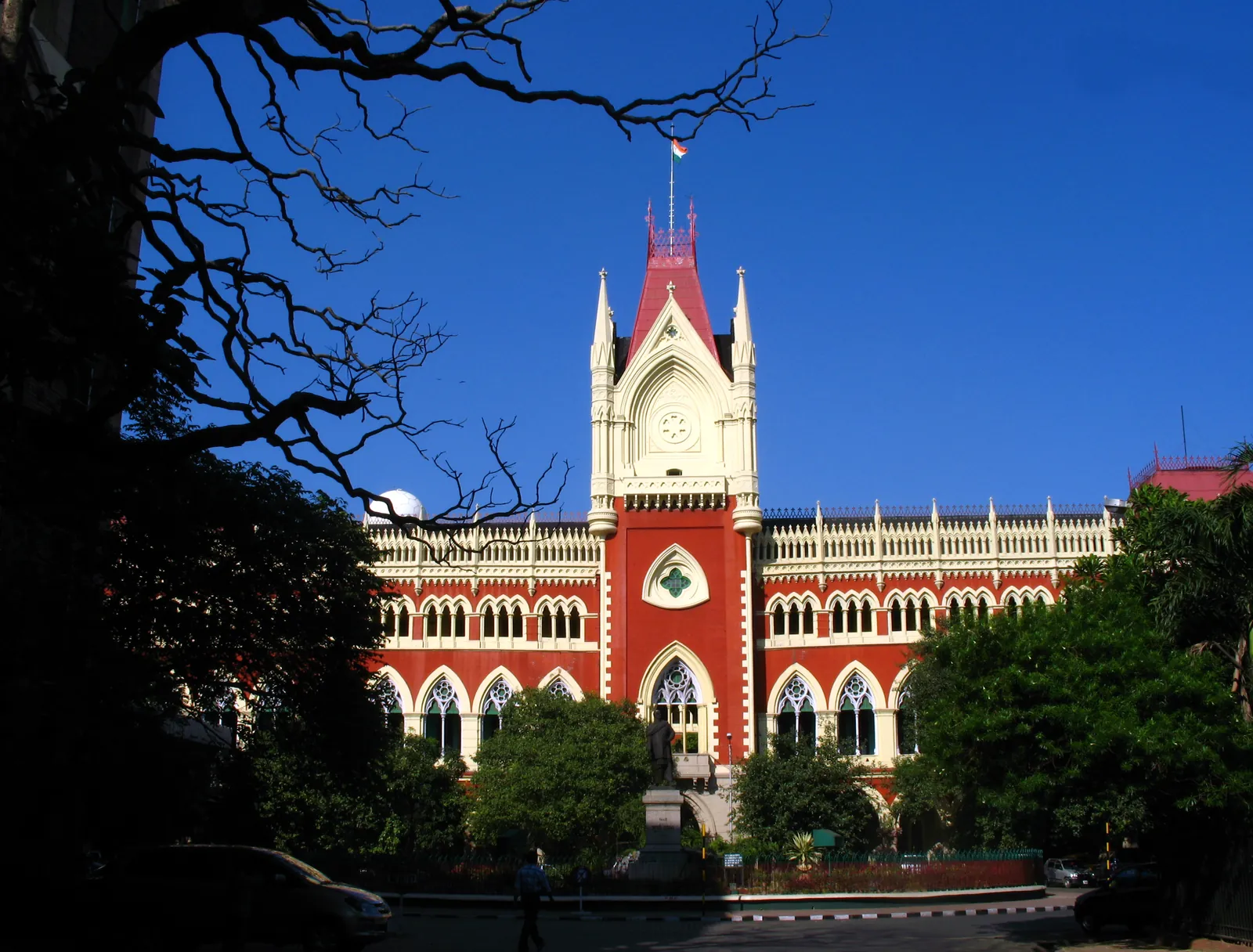The red and white Gothic facade of the Calcutta High Court with a tall central tower, pointed arches and a flag, framed by bare branches against a deep blue sky