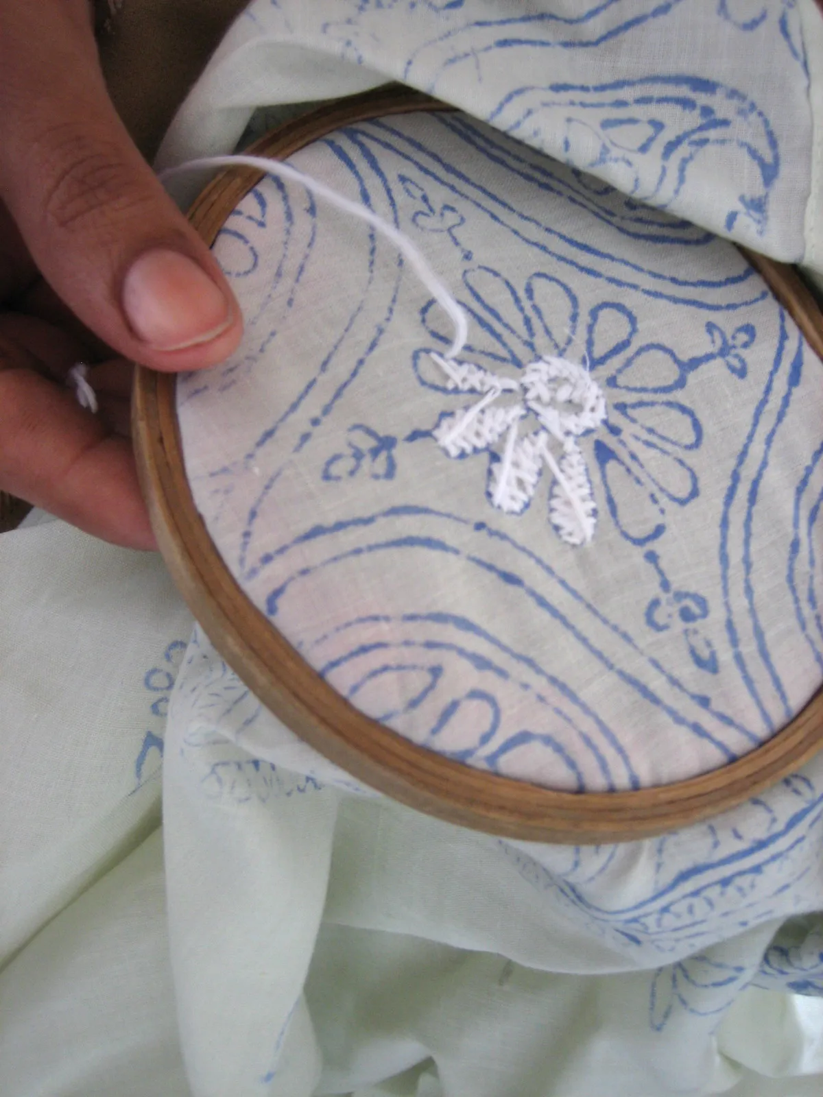 An embroiderer's hand holding a wooden hoop showing the back of pale cotton cloth, where white criss-cross stitches fill the petals of a flower printed in blue