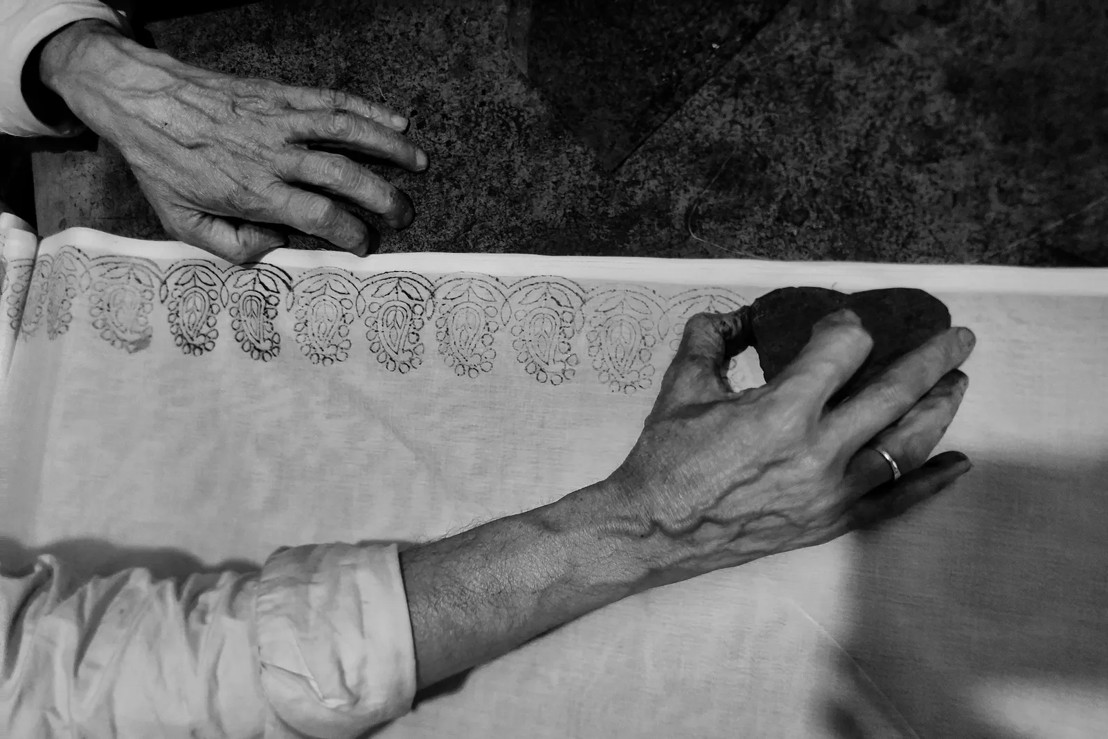 Black and white photo of an artisan's hands pressing a carved wooden block onto a length of white cloth, leaving a row of printed paisley motifs along the edge