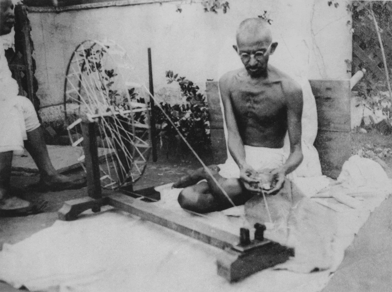 Black and white photograph of Mahatma Gandhi seated on the ground in a white dhoti, spinning yarn on a large wooden charkha with a spoked wheel