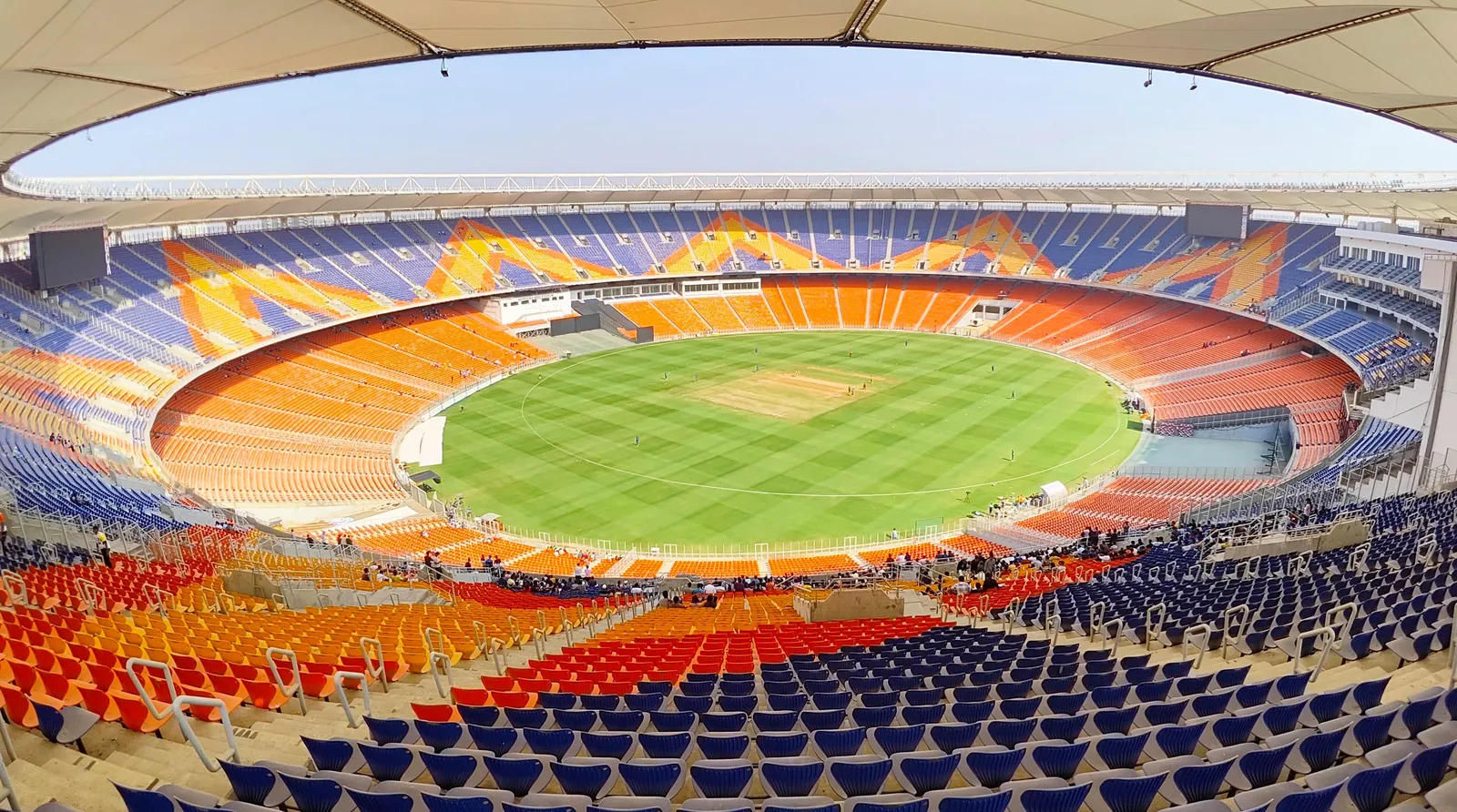 Wide view from the upper tier of the Narendra Modi Stadium in Ahmedabad, showing orange and blue seats curving around a green outfield and the pale pitch at the centre