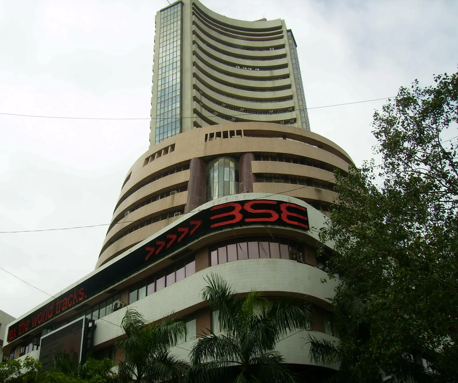 Curved tower of the Bombay Stock Exchange at Phiroze Jeejeebhoy Towers in Mumbai, with a red LED BSE ticker sign above the entrance and palm trees in front