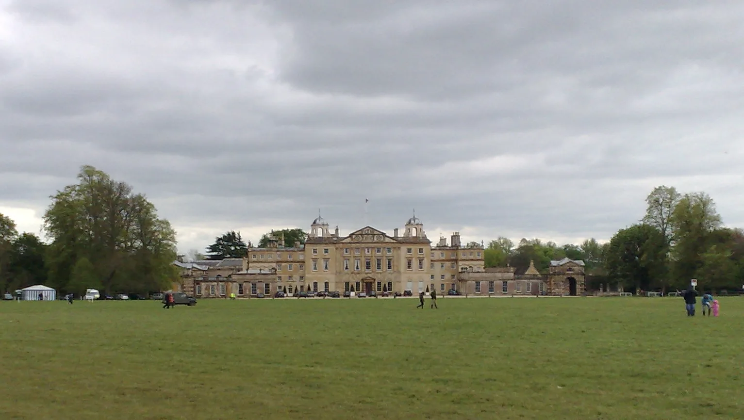 Badminton House, a large pale stone country mansion with two domed cupolas, seen across a wide green lawn under a grey sky in Gloucestershire