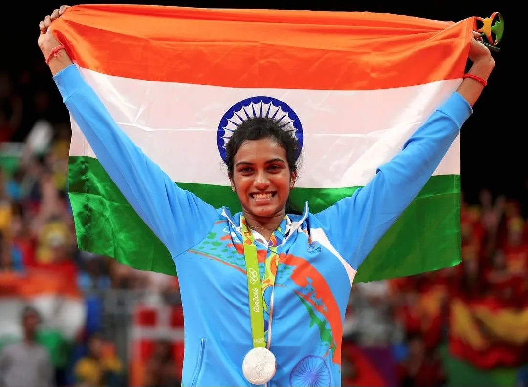 P. V. Sindhu smiling with her arms raised, holding the Indian tricolour flag behind her head and wearing an Olympic silver medal from Rio 2016