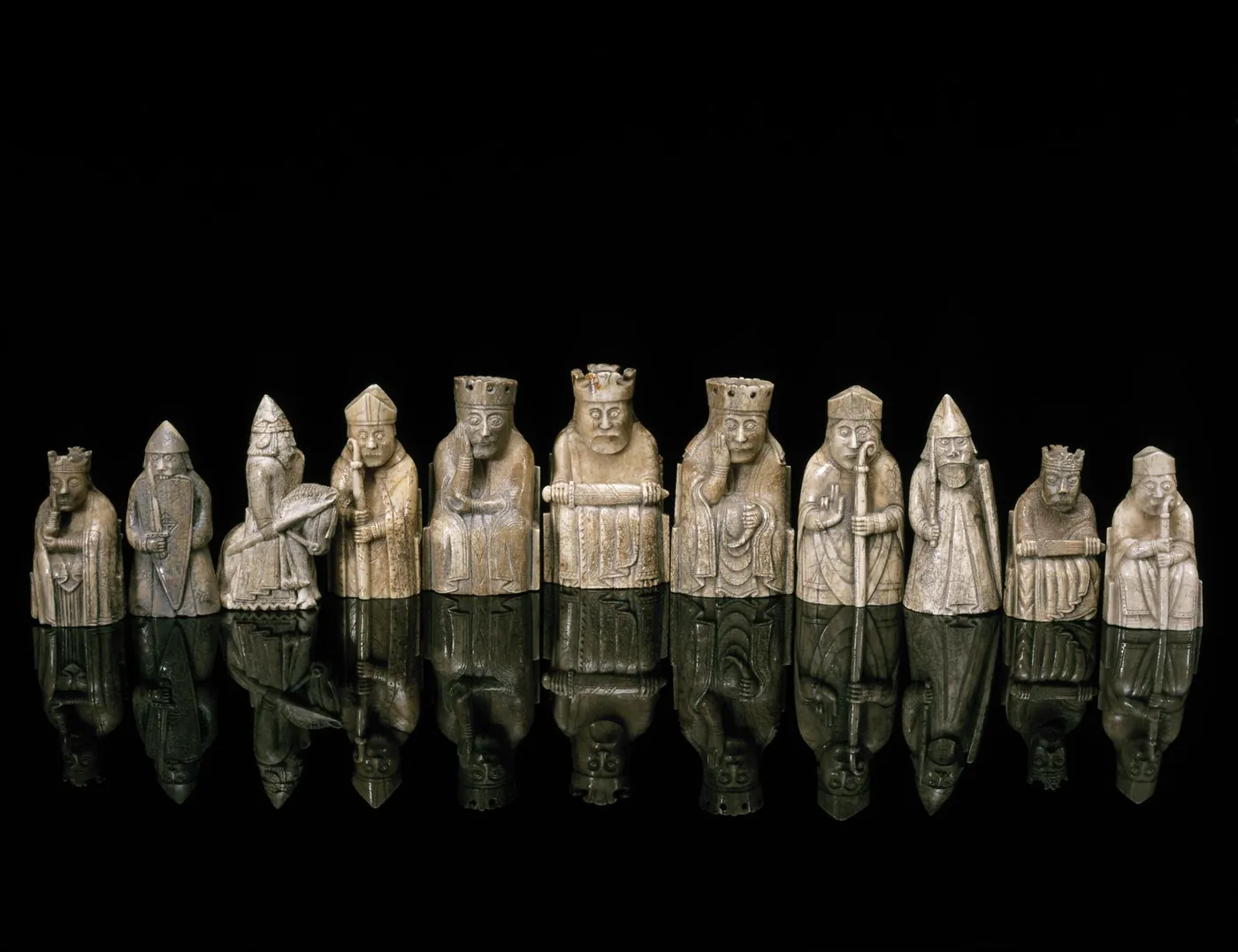 A row of carved walrus ivory chess pieces against a black background, including seated kings and queens, bishops with mitres and croziers, and helmeted warders holding shields
