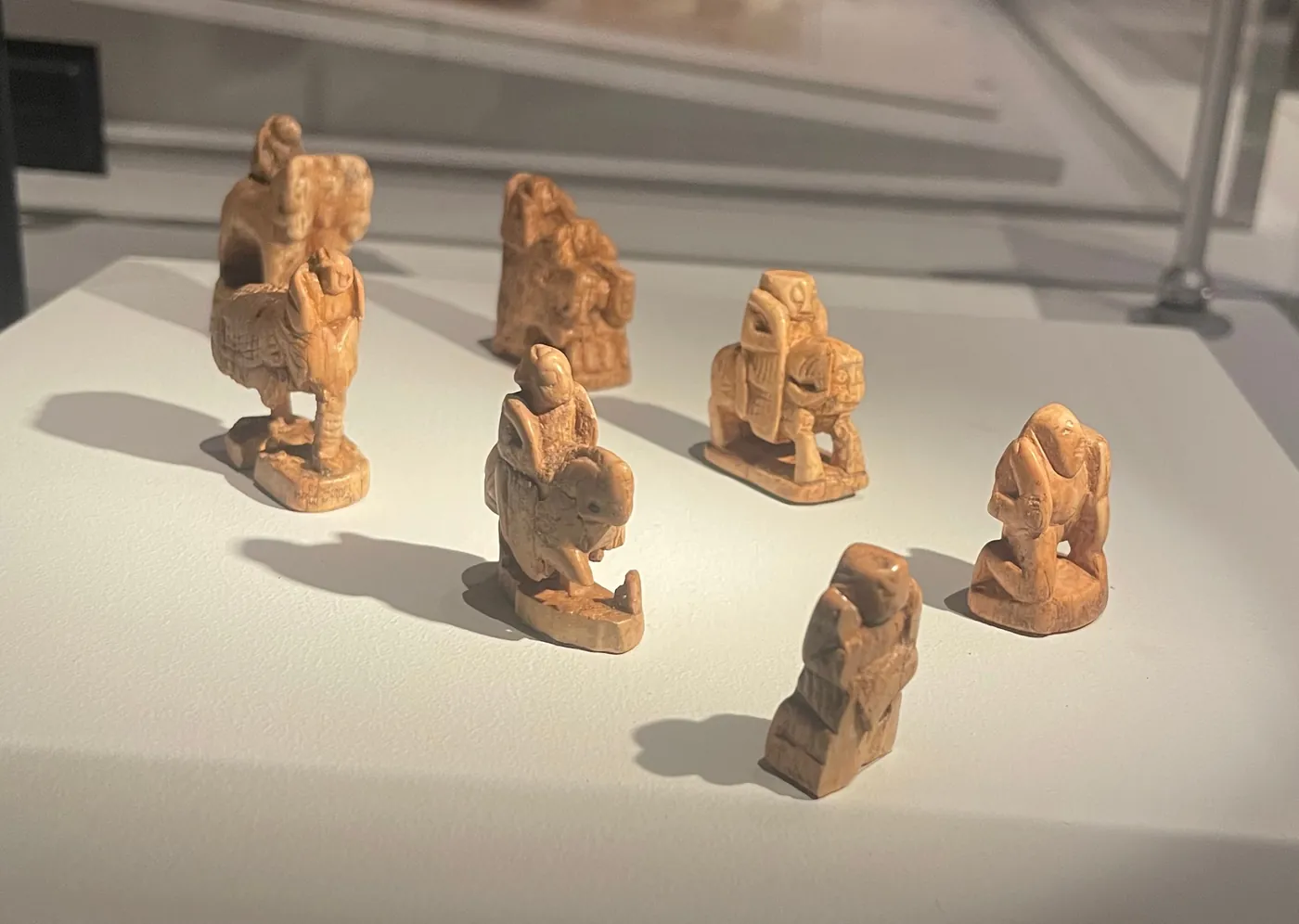 Seven small carved ivory chess pieces on a white museum plinth, including riders on horses and an elephant, worn and yellowed with age
