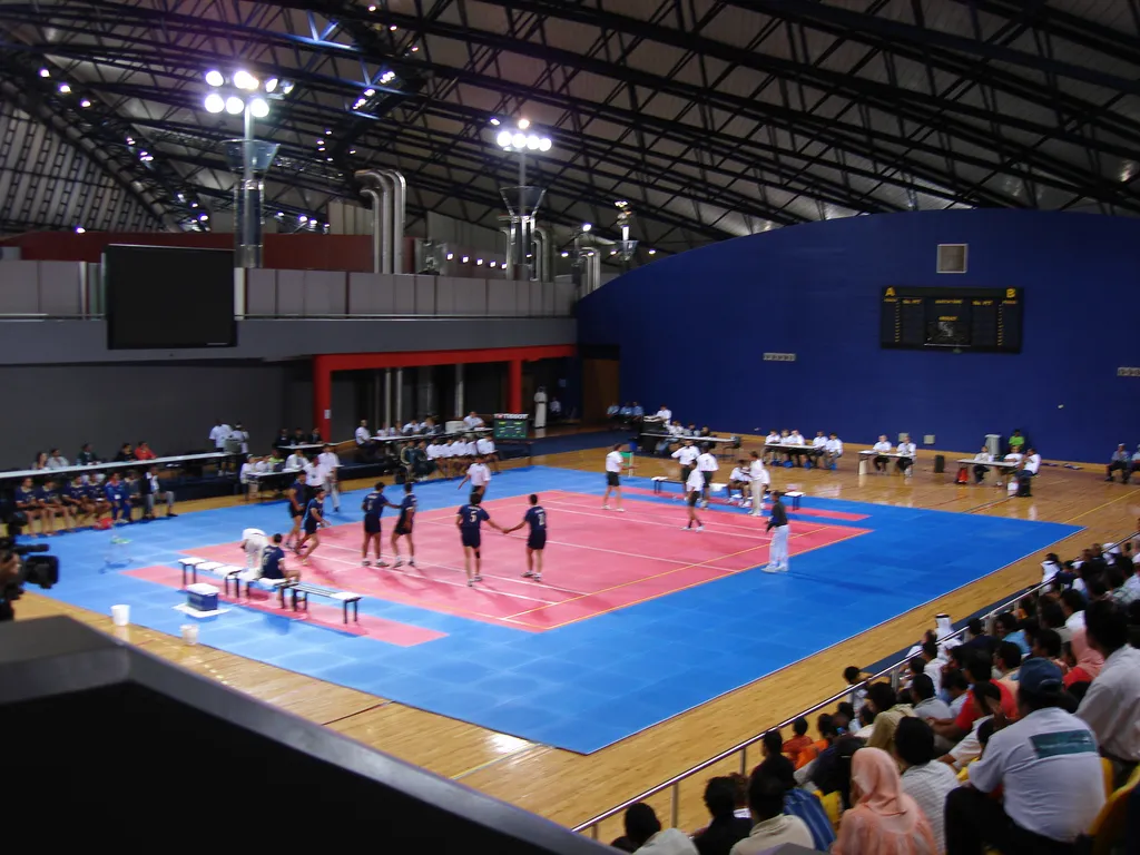 Indoor kabaddi match in Doha, with a team in dark blue shirts lined up on a red court inside a blue surround while officials and spectators watch from the sides