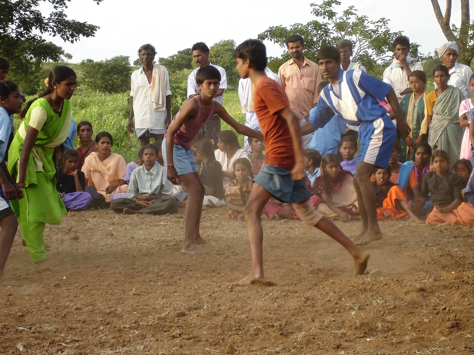 Barefoot young players face off on a dusty village kabaddi ground in India while a crowd of villagers, women in saris and children sit and stand around the edge watching