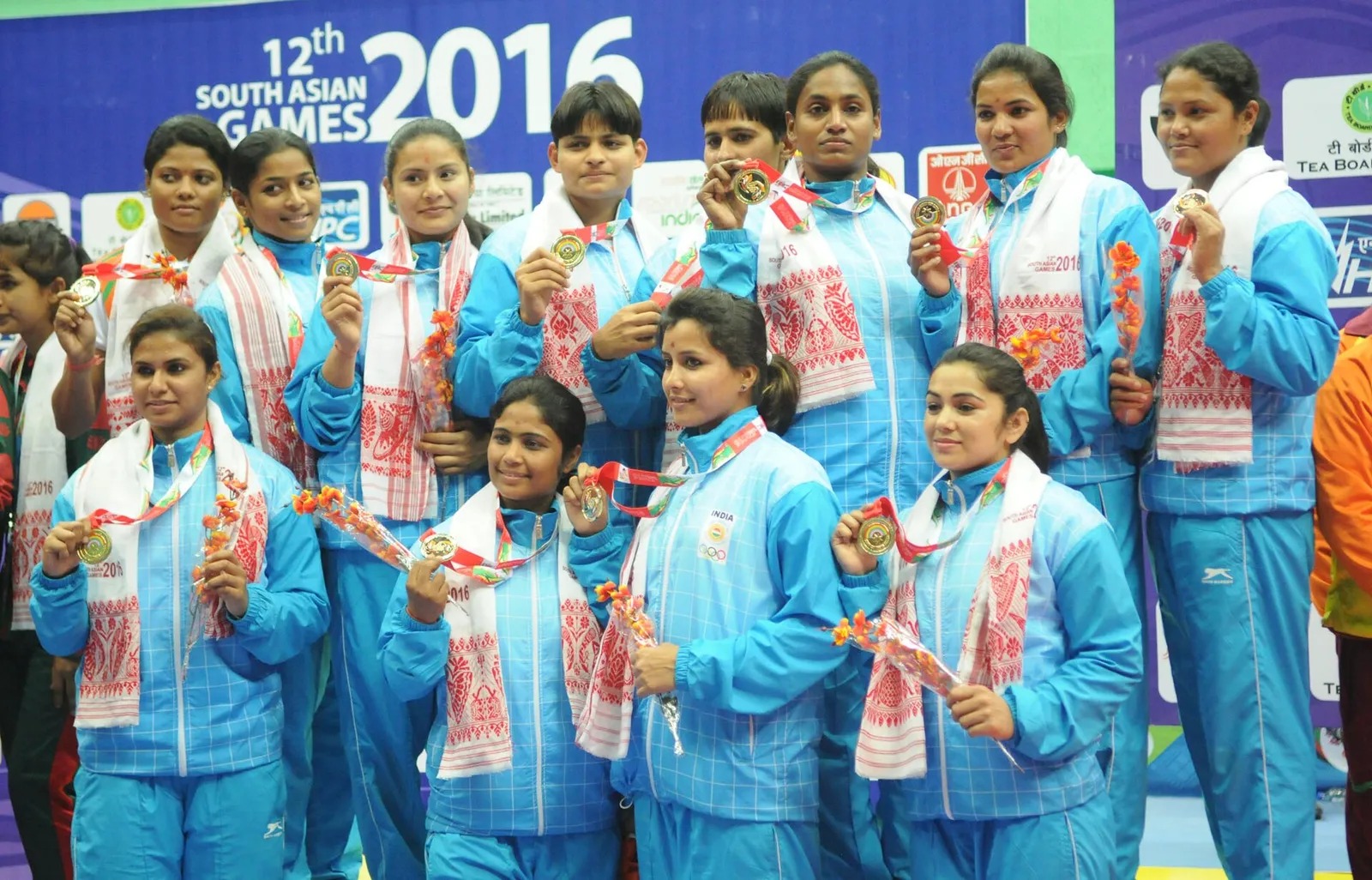 Members of the Indian women's kabaddi team in light blue tracksuits and white-and-red gamosa scarves holding up their gold medals on the podium at the 12th South Asian Games in 2016