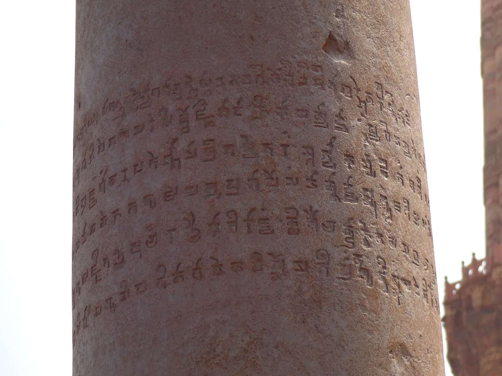 Close-up of the reddish brown surface of the iron pillar, covered in six lines of finely cut Gupta-era Brahmi letters, with the carved red sandstone of the Qutub Minar visible at the right edge