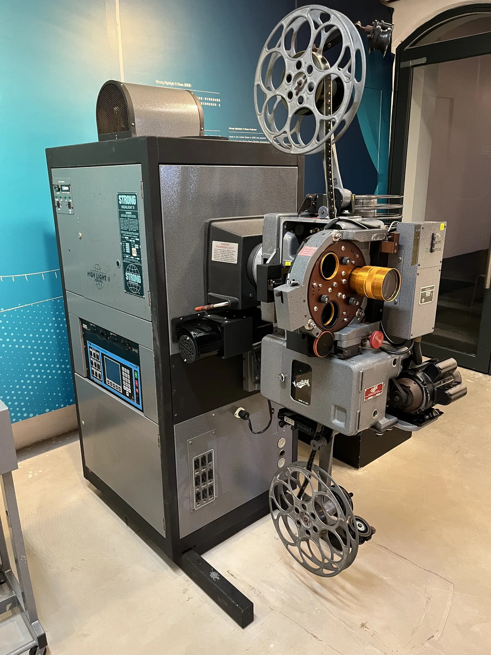 Two large 35mm film projectors standing side by side in a dim projection booth, with film reels mounted on top