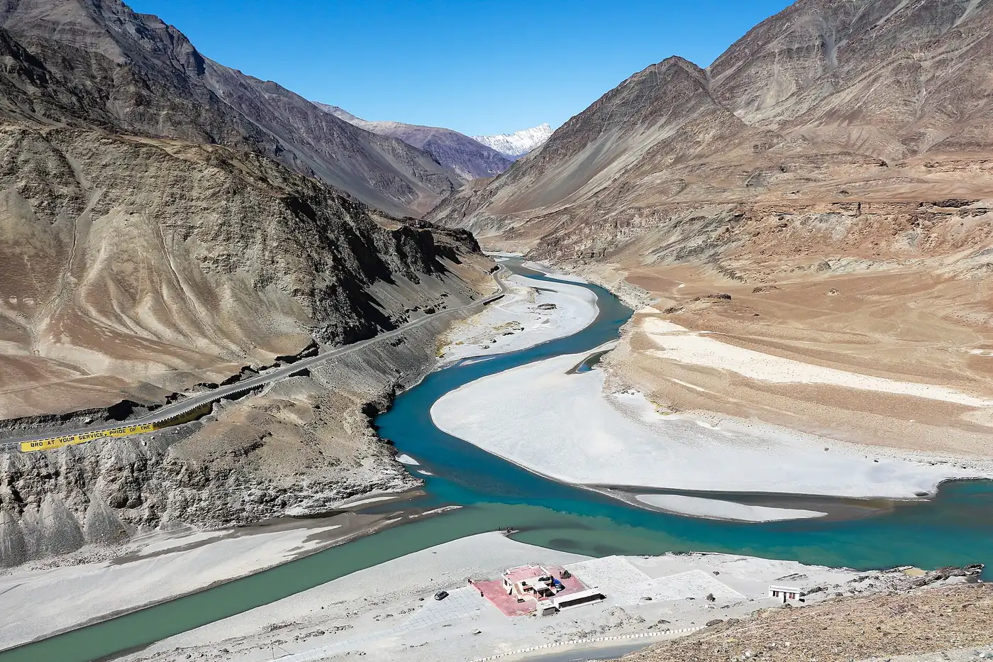 The turquoise Zanskar river meeting the greyer Indus in a wide valley of bare brown mountains in Ladakh, with a highway cut into the slope and snow peaks in the distance