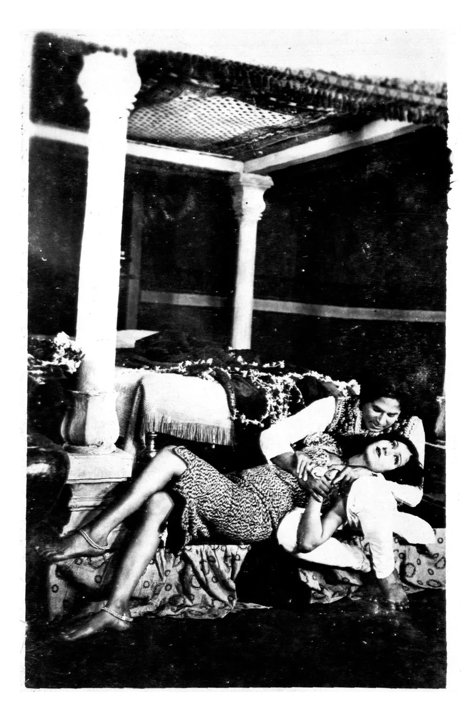 Black and white still from the 1931 film Alam Ara showing actors Master Vithal and Zubeida reclining together on a decorated bed under a canopy
