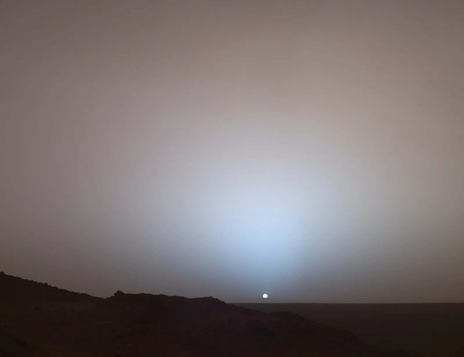 Sunset on Mars seen from Gusev crater: a small white Sun near a dark horizon, surrounded by a pale blue glow fading into a brownish grey sky