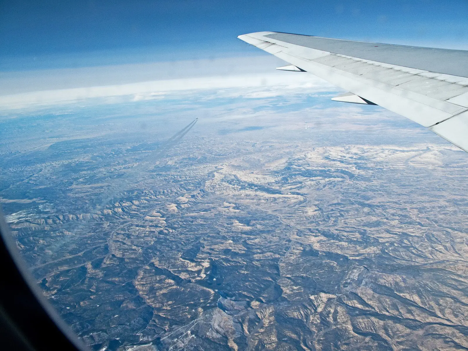 View from an airliner window at cruising altitude, with the white wing on the right and another jet leaving a contrail as it crosses far below over snow-dusted mountains